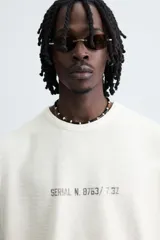 Camiseta sin mangas (manga sisa) de punto color crema, corte relaxed fit, con cuello redondo y acabados irregulares en las sisas. Presenta un texto estampado en el pecho que dice "SERIAL N. 8763/ 7.3Z".