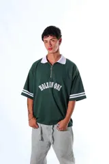 Chomba de piqué color verde con cuello polo blanco y cierre frontal parcial. Presenta un diseño holgado con mangas cortas que incluyen rayas blancas en los puños y un aplique frontal con el texto 'HOLE IN ONE' en color blanco.