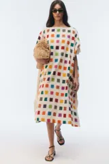 Kaftan amplio de largo midi y escote redondo, con mangas cortas caídas, aberturas laterales y ruedo asimétrico en puntas. Confeccionado en algodón, presenta un estampado de pequeños cuadrados multicolores sobre base blanca.