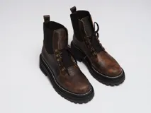 Borcego de gamuza color chocolate con paneles laterales elásticos negros y suela track dentada. Presenta sistema de cordones con ganchos metálicos y tirador en la parte trasera.