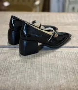 Zapato mocasín de charol negro, con punta fina, tira sobre el empeine y hebilla metálica.