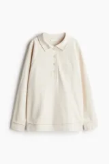 Buzo de tejido liviano color crema, de corte holgado, con cuello camisero, tapeta de botones y mangas raglán. Presenta un texto bordado en el pecho y bajo acanalado con ribete desflecado.