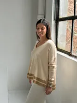 Sweater de punto color crudo con cuello en V, mangas largas y diseño holgado. Presenta detalles de franjas horizontales en color beige en los puños y en el ruedo inferior.