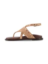 Sandalia plana de cuero gamuza color tostado, con tiras cruzadas estilo ojota adornadas con tachas metálicas doradas y tira ajustable al tobillo con hebilla.