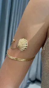 Brazalete de brazo abierto en tono dorado, con diseño de doble banda texturizada y un dije central en forma de caracol marino.