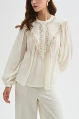 Blusa de algodón color beige, con cuello redondo y cierre frontal con botones. Presenta delicados bordados decorativos en tono tostado sobre el pecho y los hombros, mangas largas abullonadas con puños ajustados y diseño holgado.