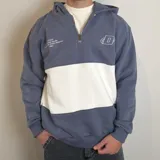 Canguro con capucha y cierre frontal parcial, de diseño color block en azul grisáceo y blanco. Presenta bordado con logo y texto en el pecho.