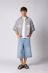 Camisa de manga corta de corte boxy, confeccionada en popelina a rayas celestes y blancas. Presenta cuello camisero clásico, cierre frontal con botones y estampado tipográfico en el pecho.