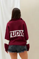 Sweater bordó con cuello alto y cierre frontal parcial, de diseño holgado con franjas blancas en las mangas y una franja blanca ancha en la espalda con la inscripción en letras bordó.