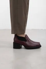 Botín asimétrico de estilo Chelsea color bordó, confeccionado en cuero vacuno, con paneles elásticos laterales negros, puntera cuadrada, suela de plataforma y tacón de bloque cuadrado.