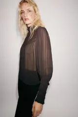 Blusa de cuello subido y manga larga, color marrón oscuro, con detalles de bordados combinados a tono en el canesú y los puños anchos. Cierre en la espalda con abertura y botones.