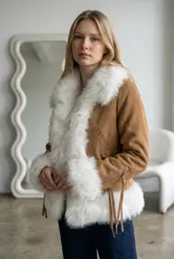Abrigo de gamuza color beige con detalles de piel sintética blanca en el cuello, puños y ruedo. Presenta cierre frontal, bolsillos laterales con cierre y flecos decorativos en las mangas.