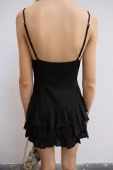 Vestido corto negro de tirantes finos ajustables y escote en V. Presenta un diseño ajustado con drapeado central y cordones cruzados laterales tipo corsé, y falda corta con tres capas de volantes escalonados.