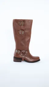 Bota de caña alta confeccionada en cuero color suela, con diseño de estilo western. Presenta tres hebillas metálicas en la parte superior de la caña y un estribo con argolla metálica en el tobillo. Cuenta con taco cuadrado de altura media.