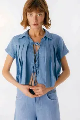 Blusa de lino color azul, de corte cropped y manga corta. Presenta cuello camisero, diseño con tablas frontales y cierre mediante lazos delgados en el centro.