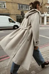 Abrigo de paño negro con capucha, cierre frontal cruzado con botones y cinturón de lazada en el mismo tejido. Presenta manga larga con trabillas en los puños y bolsillos laterales.