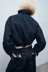 Cazadora de mezclilla azul oscuro, con cuello alto, manga larga, bolsillos ribeteados en el delantero y cierre frontal con cremallera y botones a presión ocultos bajo una solapa. Presenta costuras con pespuntes en contraste.