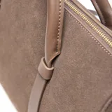 Cartera tipo baúl compacta, color marrón, con material exterior de símil gamuza combinado con PU liso. Cuenta con doble asa corta rígida, correa larga regulable y desmontable, y herrajes metálicos dorados. El compartimento principal tiene cierre superior y un bolsillo interno con cierre. Incluye detalle frontal con logo XL metalizado.