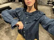 Camisa blanca de textura rugosa con volados en el pecho y hombros, cuello redondo con lazos de ajuste, cierre frontal con botones y mangas largas con puños con volados.