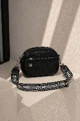 Cartera bandolera negra de textura granulada, con bolsillo frontal con cierre y herrajes metálicos plateados. Incluye correa ajustable de tela con diseño bordado en blanco y negro.