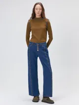 Jeans de corte ancho (wide leg) y tiro medio, confeccionados en denim de algodón negro con costuras contrastantes en color ocre. Presenta múltiples botones metálicos frontales y tachas metálicas aplicadas a lo largo de los laterales y la pretina.