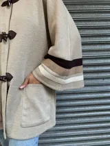 Saco color beige de lana con cierre frontal con broches tipo tanca de cuero. Presenta mangas anchas con detalle de rayas horizontales en marrón y beige.