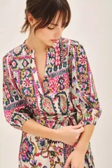 Vestido corto de cuello redondo con escote en V, mangas 3/4 con puños elásticos y lazo ajustable en la cintura. Presenta un estampado multicolor con motivos geométricos y florales.