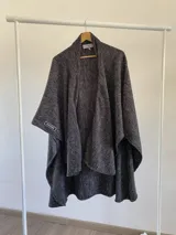 Ruana abierta de tejido con estructura, color negro con patrón de espiga en gris. Presenta un diseño envolvente con caída fluida y etiqueta de marca en el borde.