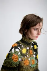 Jersey de punto con cuello subido y manga larga. Presenta un diseño de rayas horizontales en tonos verde y negro, con un panel superior decorado con calados y aplicaciones de flores tejidas en crochet de colores contrastantes.