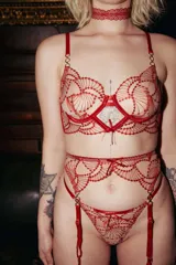 Conjunto de lencería de tres piezas en color rojo, confeccionado en tela transparente con bordados ornamentales. Incluye soutiene con aro y tirantes ajustables, bombacha colaless y portaligas a juego con detalles de argollas y apliques.