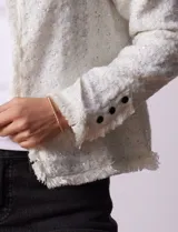 Chaqueta de tejido tweed color blanco con destellos brillantes, de corte recto y estructurado. Presenta cuello a la base, abertura frontal y puños decorados con botones en contraste. Los bordes del cuello, la cartera frontal y el ruedo cuentan con una terminación deshilachada.