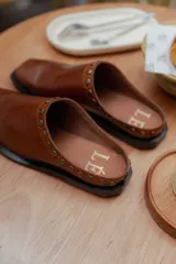 Mules de cuero color suela, con diseño abierto y tira frontal decorada con tachas metálicas circulares. Presentan punta cuadrada y suela plana.