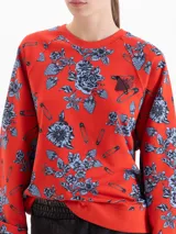 Buzo de algodón con cuello redondo y mangas largas, estampado con diseño de flores y alfileres de gancho sobre fondo rojo. Presenta puños y ruedo en tejido elástico al tono.