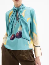 Camisa de manga larga color celeste con estampado de lunas amarillas y cerezas rojas. Presenta cuello con lazo anudado y ribetes en azul marino en el cuello, puños y cartera frontal.