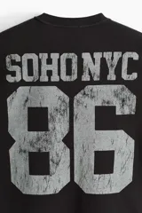 Remera de algodón en punto pesado color negro, de corte holgado con hombros caídos, cuello redondo acanalado y estampa frontal estilo universitario con el texto 'SOHO NYC 86'.