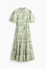 Vestido largo de algodón color blanco con estampado botánico en tonos verdes. Presenta cuello mao, cierre frontal con botones ocultos, mangas cortas abullonadas y falda de corte escalonado con volados. Incluye un cinturón fino para ajustar en la cintura.