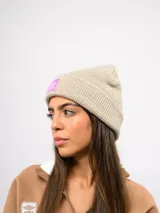 Gorro de lana tipo beanie de tejido acanalado, color beige melange, con vuelta y parche cuadrado en color fucsia con el logo de Umbro.