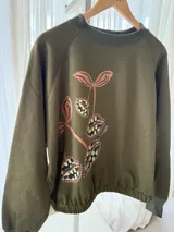 Buzo cropped de algodón color verde militar, con mangas ranglan y diseño estampado en el frente con motivos de besos y moños. Cuenta con elástico holgado en la cintura y los puños.