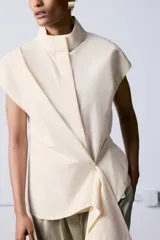 Camisa sin mangas confeccionada en algodón, con cuello subido y diseño cruzado con detalle de fajín asimétrico lateral. Cierre frontal con botones ocultos.