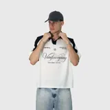 Polo de corte oversize con diseño bicolor, cuerpo en color blanco y mangas raglán en color negro. Presenta cuello tipo polo en negro y estampado gráfico frontal con tipografía cursiva y detalles de la marca.