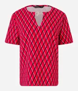 Blusa de crepé color rojo con estampado geométrico de rombos en negro y blanco. Tiene cuello redondo con abertura en forma de gota y detalle metálico, mangas cortas y corte recto.