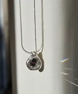 Collar plateado con dije circular con piedra facetada transparente. Cadena fina de eslabones. Disponible en plata 950 o con baño de oro 24K.