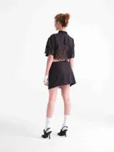 Camisa crop negra de tencel, con mangas cortas y detalles de encaje en el cuello, la botonera frontal, el dobladillo inferior y las mangas. Cuenta con tiras ajustables que se atan alrededor de la cintura.