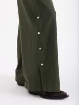 Pantalón de corte recto y silueta holgada, confeccionado en tejido rústico de algodón color verde militar. Presenta cintura con elástico interno y detalles de broches metálicos a presión en los laterales.