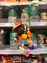 Llavero con figura de oso de peluche marrón, vestido con delantal a cuadros rojos y blancos y sombrero de paja con flor roja. El oso sostiene un objeto pequeño y está adornado con un collar de cuentas de colores y dijes con letras que forman una palabra.