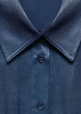 Vestido midi de estilo camisero en tejido satinado fluido, color azul. Presenta cuello camisero, cierre frontal con botones, mangas cortas con dobladillo y dos bolsillos de parche en el pecho. Incluye un lazo ajustable en la cintura para entallar la silueta.