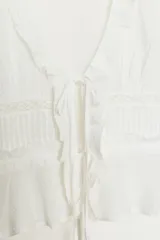 Camisa de gasa color blanco con escote en pico, cierre frontal con lazadas y detalles de volantes. Presenta mangas largas con puños elásticos y terminación en volante, además de bordados a tono.