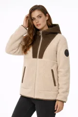 Campera de corderito sintético en color beige, con paneles en color marrón en la zona del pecho, capucha y bolsillos. Presenta cierre frontal completo y un parche circular decorativo en la manga.