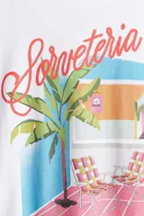 Remera blanca de algodón con cuello redondo y hombros caídos, con un estampado gráfico que muestra una escena de playa o terraza con una palmera, sillas de lona a cuadros y el texto "Sorveteria Rio de Janeiro".