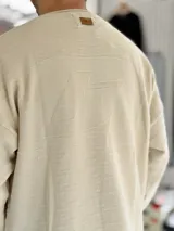 Sweater de punto liviano color beige, con textura marcada, cuello redondo y mangas largas de corte relajado, con pequeño estampado tipográfico negro en el lateral inferior.